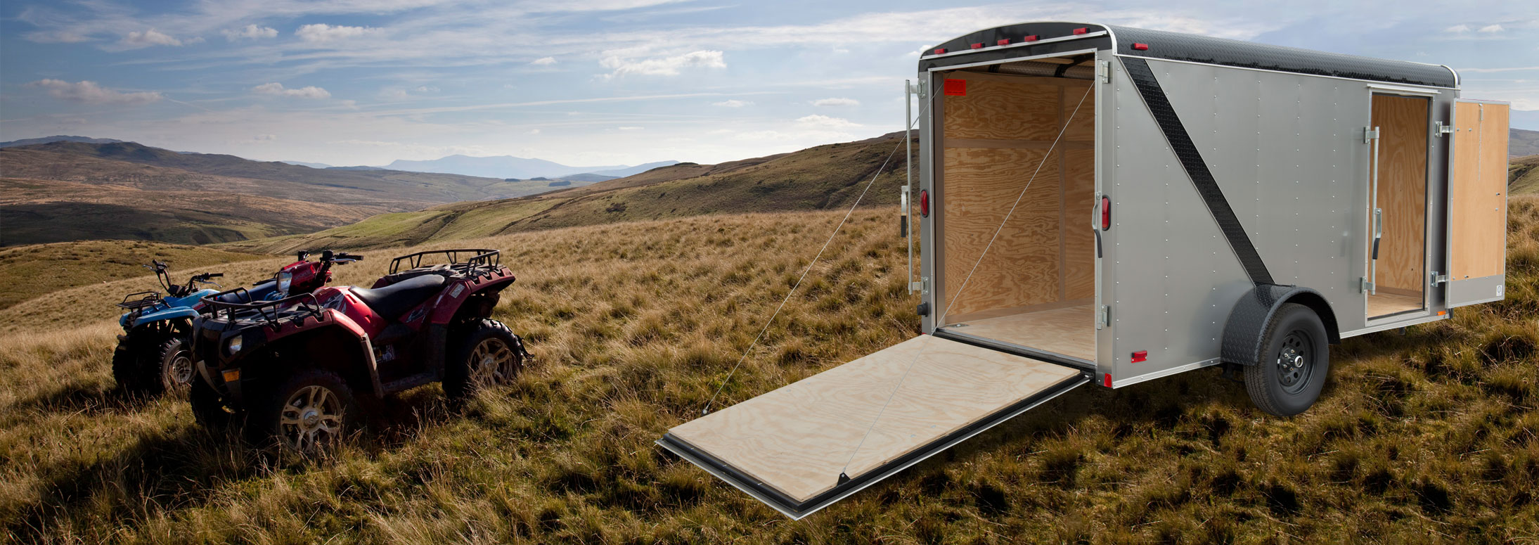 Two four wheelers in a field next to a cargo trailer